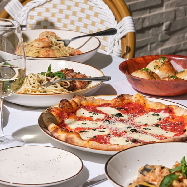A table laden with Italian dishes: pizza, pasta with shrimp, chicken pasta, and garlic knots.