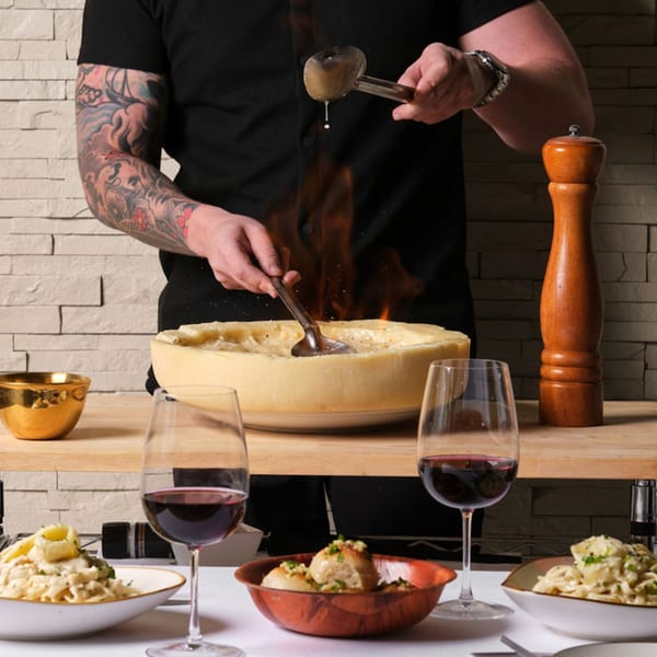 Chef flambéing pasta in a large cheese wheel, with wine glasses and pasta dishes on a table.