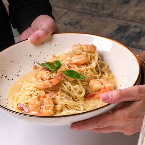 Hands holding a white bowl of shrimp scampi with linguine pasta and basil garnish.