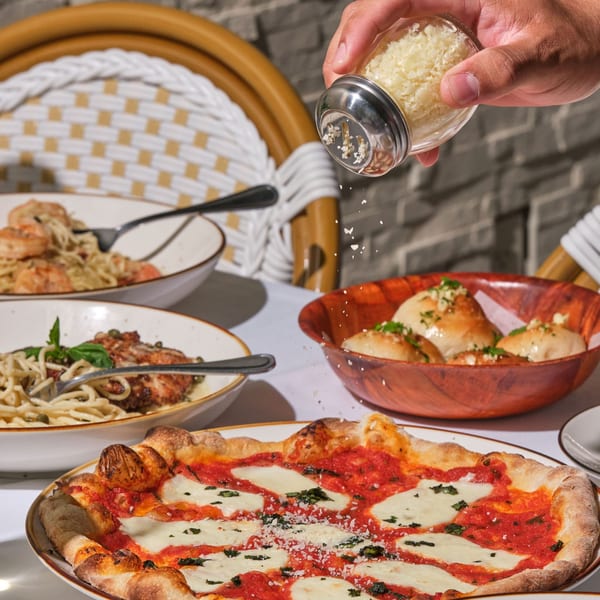 Hand sprinkling Parmesan cheese over a Margherita pizza, with pasta dishes and garlic knots on a white table.