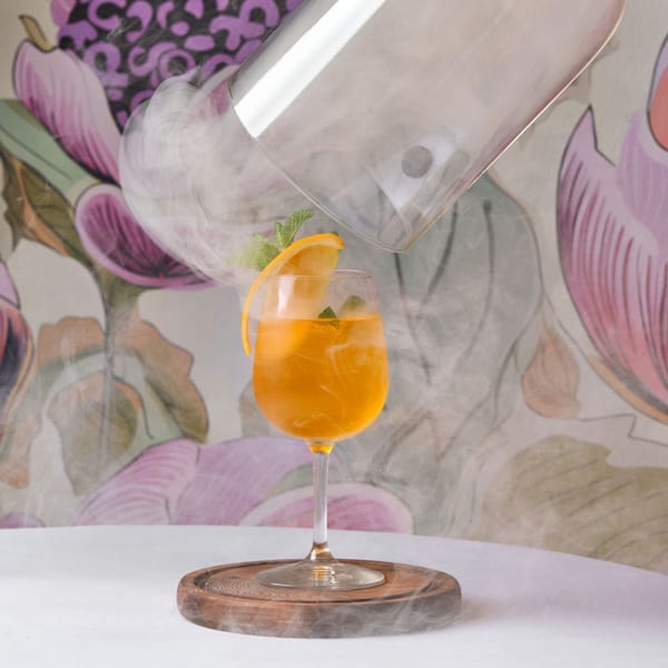 A smoked orange cocktail with mint and an orange peel garnish on a wooden coaster.