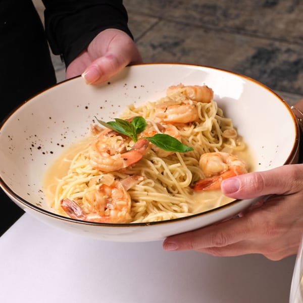 Hands holding a white bowl of shrimp scampi with linguine and basil garnish.