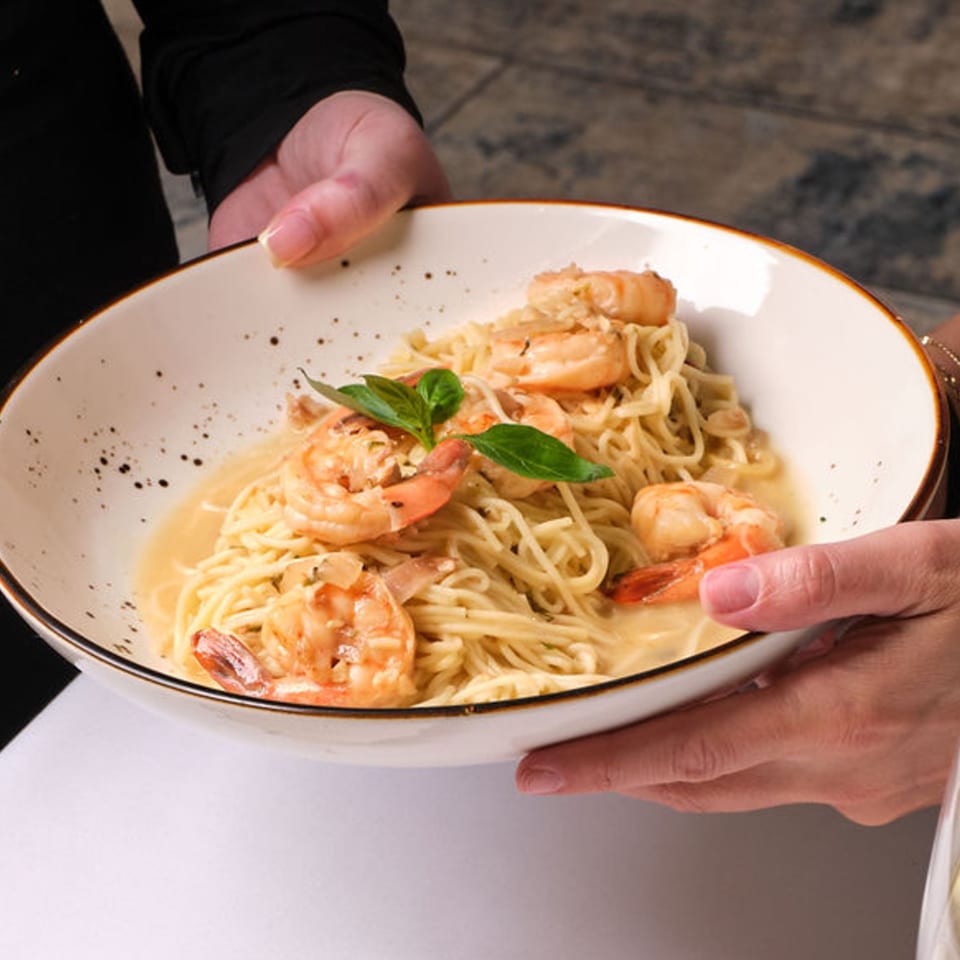 Hands holding a white bowl of shrimp scampi with linguine and basil garnish.