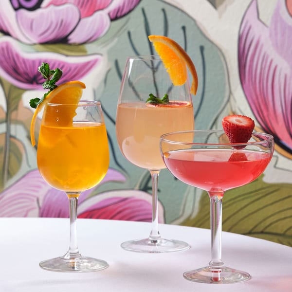 Three colorful cocktails with fruit garnishes on a white table in front of a floral wallpaper.