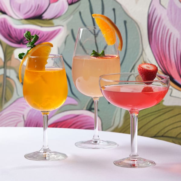 Three colorful cocktails, garnished with orange slices, mint, and a strawberry, on a white table.