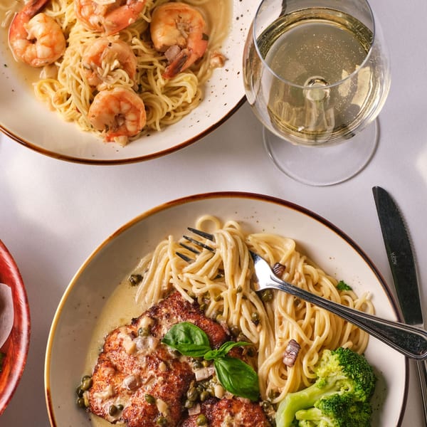 Two plates of pasta, one with shrimp, the other with chicken, broccoli, and a glass of white wine.
