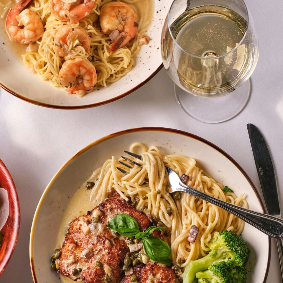 Two plates of pasta, one with shrimp, the other with chicken, broccoli, and a glass of white wine.