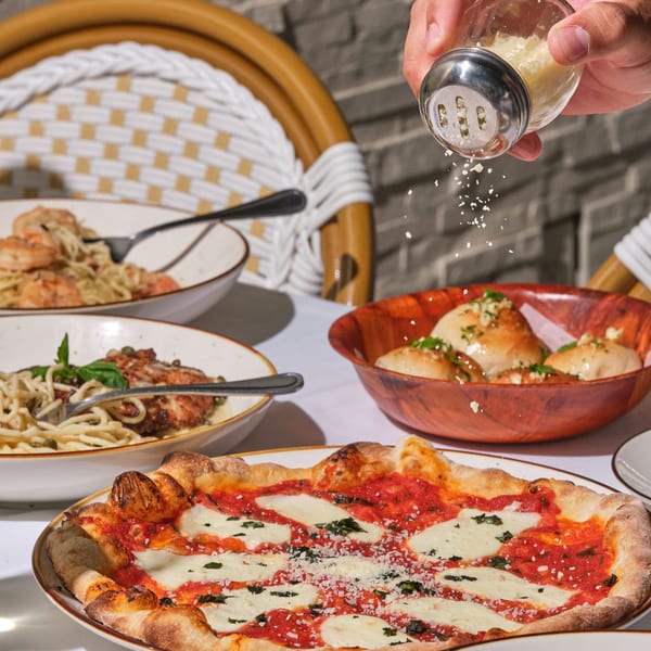 Hand sprinkling parmesan cheese over a margherita pizza, pasta dishes, and garlic knots on a table.